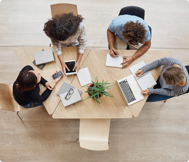 A bird's-eye view of four office workers seated at their desks, each engrossed in their electronic devices. Laptops, tablets, and smartphones are visible on the desks, with papers and notebooks scattered around. The workers appear focused, typing or scrolling intently on their screens.